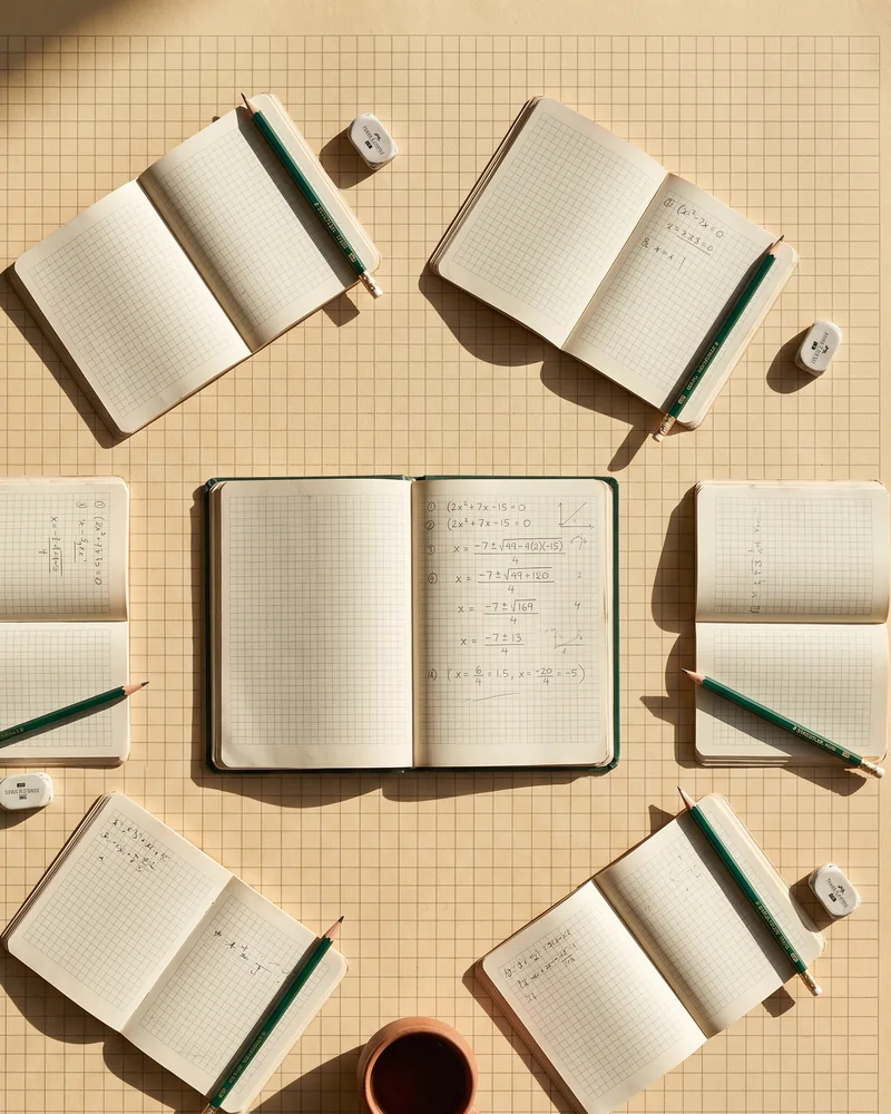Several exercise books arranged around one central open notebook on squared paper, each with its own pencil and eraser