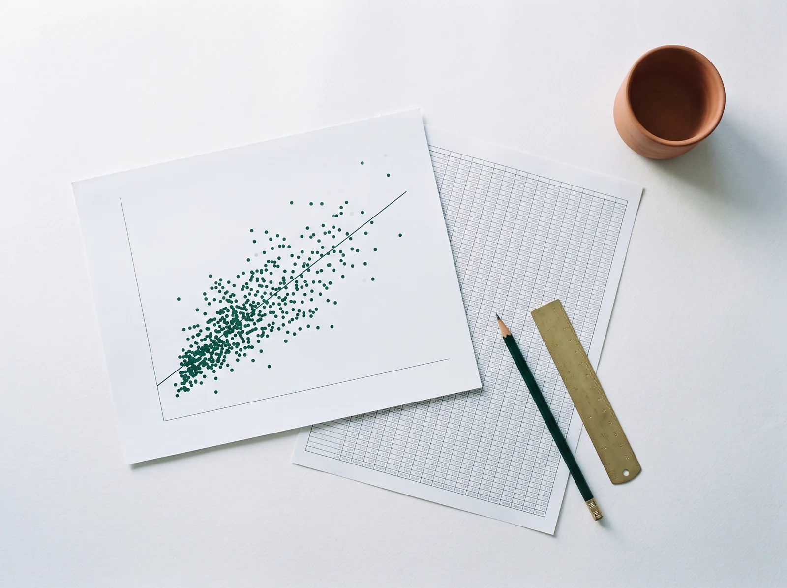 A printed scatter plot with a fitted trend line beside a table of figures, with a pencil and a brass ruler resting across them